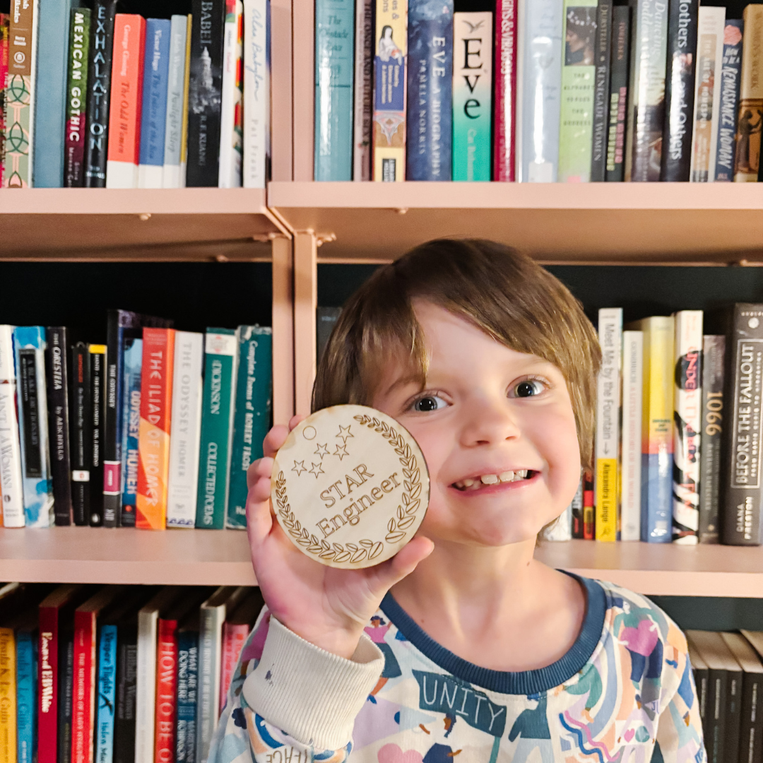 Brontë stands in front of a bookcase. She is smiling and holding a wood medal that is engraved with the words, STAR Engineer. 